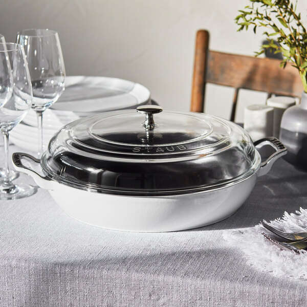 A white Staub enameled cast iron pot with a glass lid on a table.