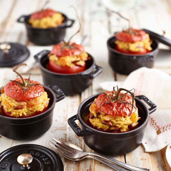 A group of black Staub enameled cast iron mini Dutch ovens filled with food with a tomato on top.