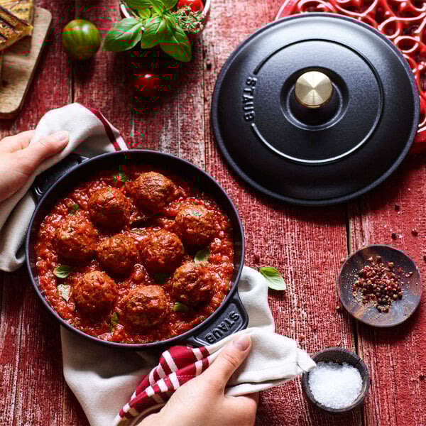 A person holding a Staub black enameled cast iron pot of meatballs with red sauce and a towel.
