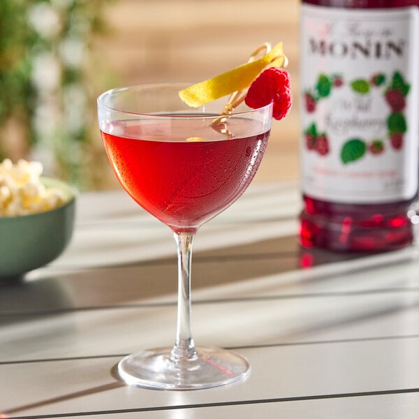 A glass of red raspberry-flavored drink garnished with a lemon twist and raspberry, with a bottle of Monin Premium Wild Raspberry Flavoring Syrup in the background.