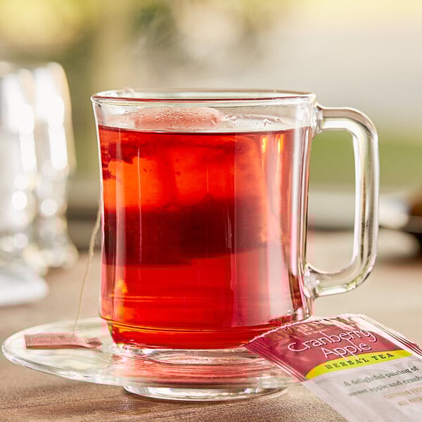 A glass mug with red liquid on a saucer with a Bigelow Cranberry Apple Tea bag.