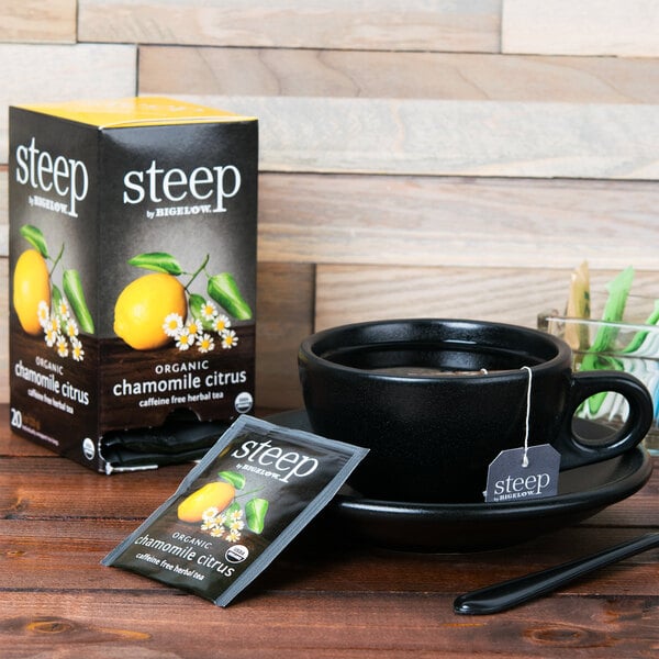 A black coffee mug with a tea bag on a black plate next to a box of Steep By Bigelow Organic Chamomile Citrus Tea.