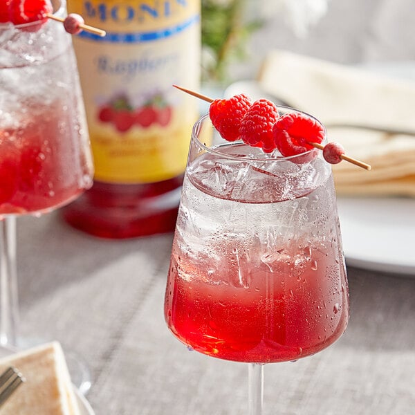 A glass of red liquid with ice and raspberries on a table with a Monin Sugar Free Raspberry Fruit Syrup bottle.