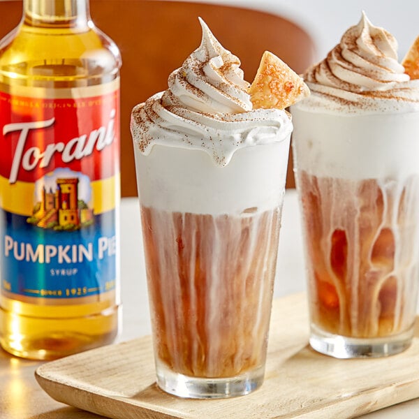 Two glasses of brown drinks with whipped cream on top and a bottle of Torani Pumpkin Pie flavoring syrup.