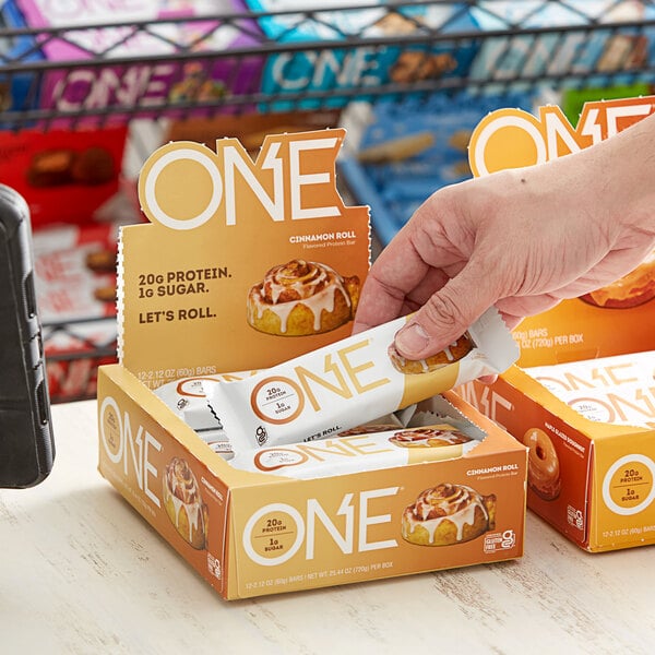 A person's hand holds a box of ONE Cinnamon Roll Protein Bars.