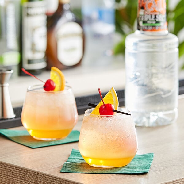 Two glasses of orange drinks garnished with cherries next to a bottle of Finest Call Triple Sec syrup.