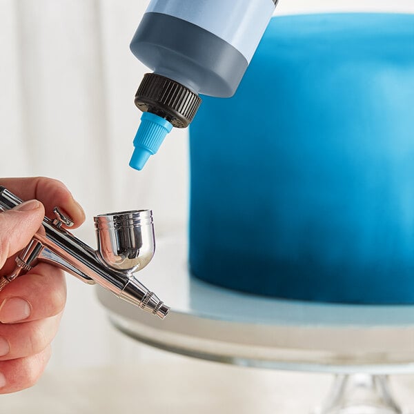 A person using a silver and blue Chefmaster airbrush to paint a cake blue.