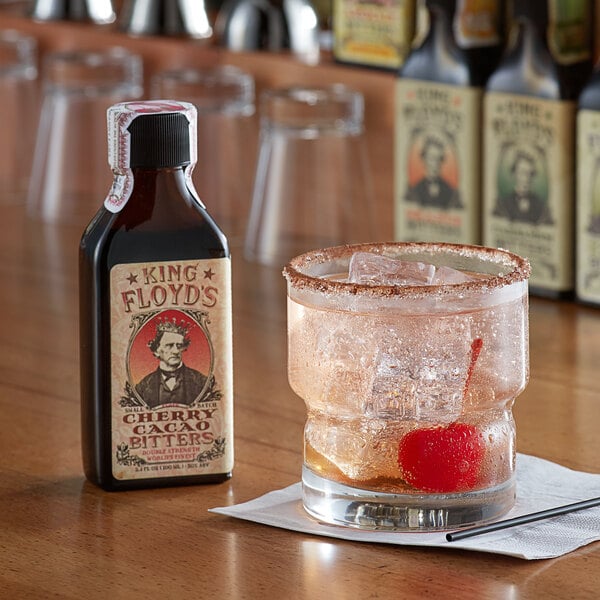 A bottle of King Floyd's Cherry Cacao Bitters on a table next to a glass with ice and a cherry.