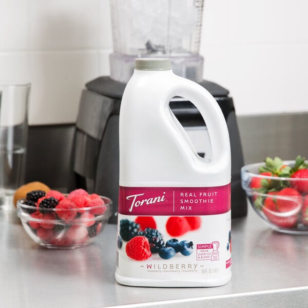 A white Torani bottle of Wildberry Fruit Smoothie Mix on a counter with bowls of berries.