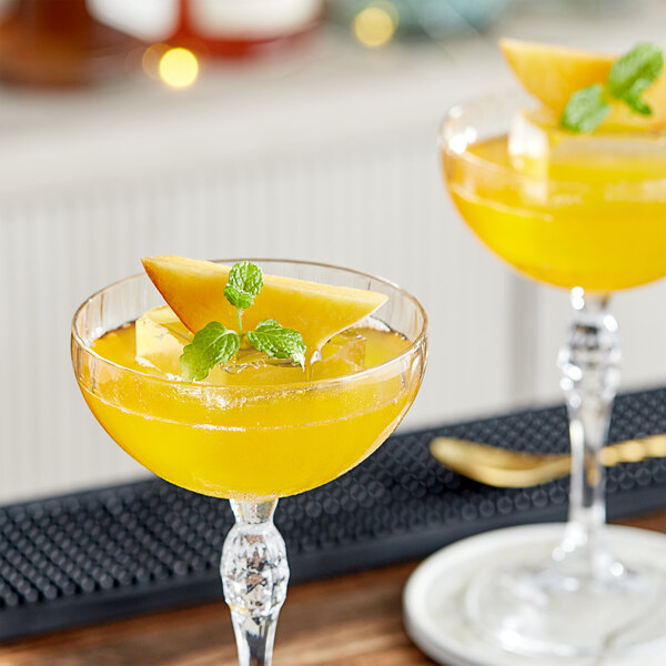 Two glasses of yellow peach cocktails garnished with mint leaves and fruit slices.