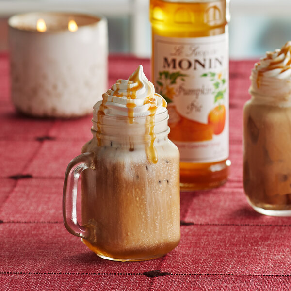 A glass mug of a drink with whipped cream and caramel syrup next to a bottle of Monin Premium Pumpkin Pie Flavoring syrup.