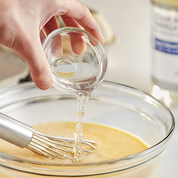 A hand pouring McCormick Culinary Clear Imitation Vanilla into a bowl of liquid on a counter in a professional kitchen.