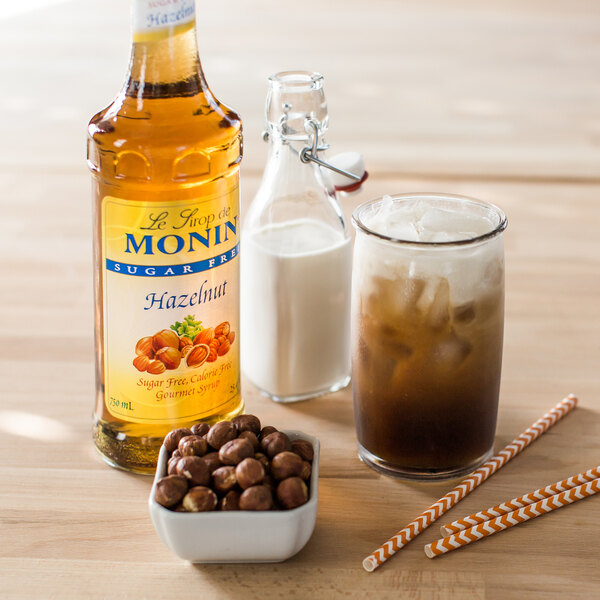 A bottle of Monin Sugar Free Hazelnut flavoring syrup next to a bowl of hazelnuts and a glass of milk.