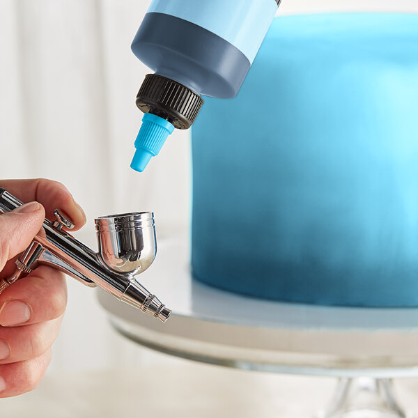 A person using a metal airbrush to paint a cake blue with Chefmaster Neon Brite Blue food coloring.