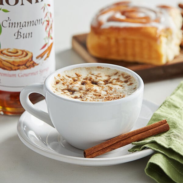 A cup of coffee with a bottle of Monin Cinnamon Bun flavoring syrup and cinnamon sticks on a table.