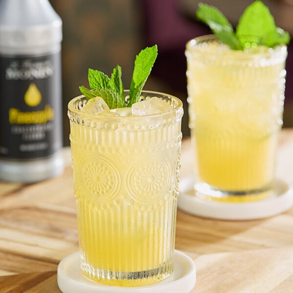 A glass of yellow pineapple-flavored beverage with ice and mint garnish, placed on a wooden surface.
