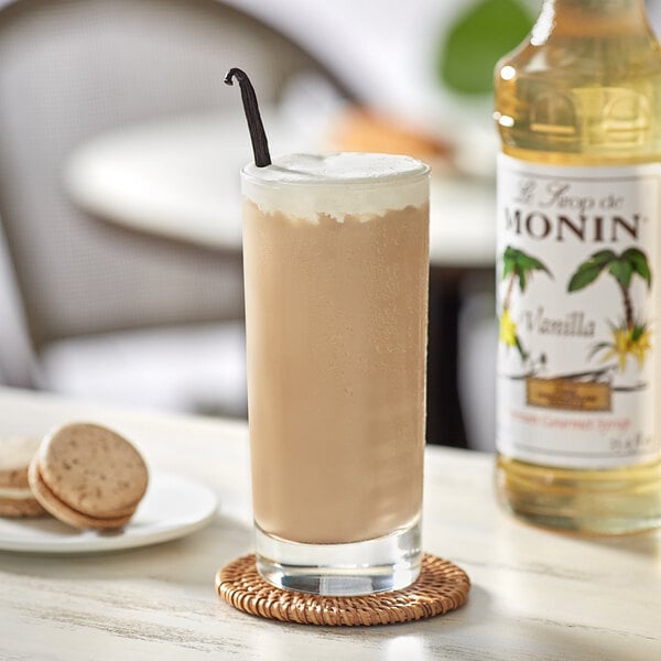 A glass of brown liquid with a straw and a bottle of Monin Premium Vanilla Flavoring Syrup.