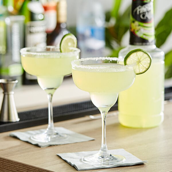 Two glasses of Finest Call margarita on a bar with lime slices.