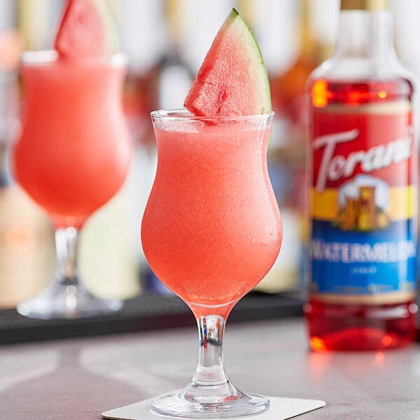 A watermelon drink in a glass with red liquid and a Torani Watermelon Flavoring Syrup bottle on a table.