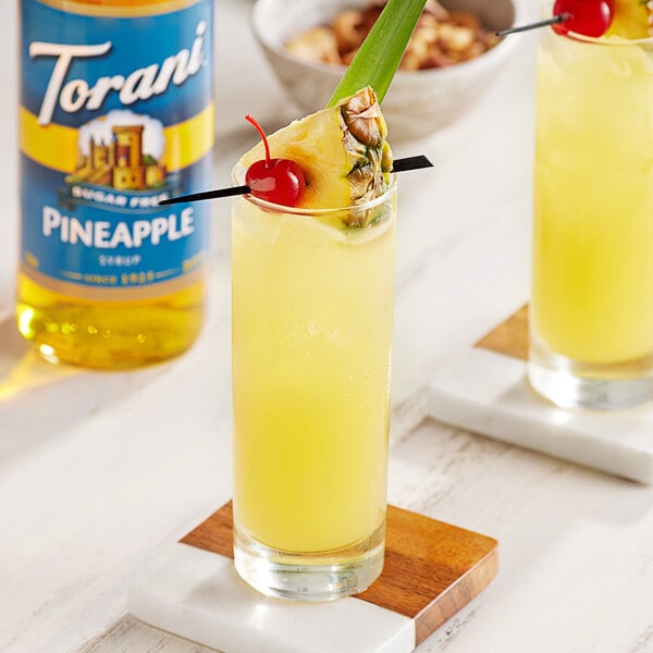 Two glasses of yellow drinks with pineapple and cherries on a table.