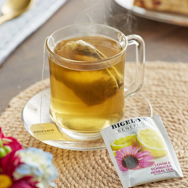 A glass mug of Bigelow Lemon and Echinacea tea with a tea bag in it on a plate with a lemon slice and a flower.