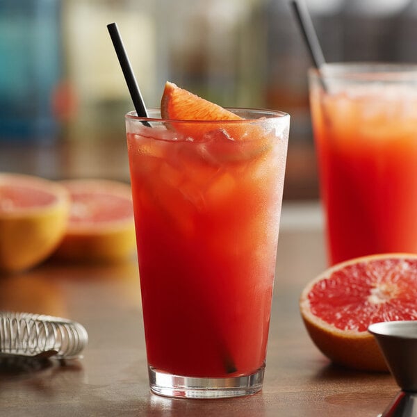 A glass of red liquid with a straw next to a half of a grapefruit on a table with a Monin Blood Orange drink.