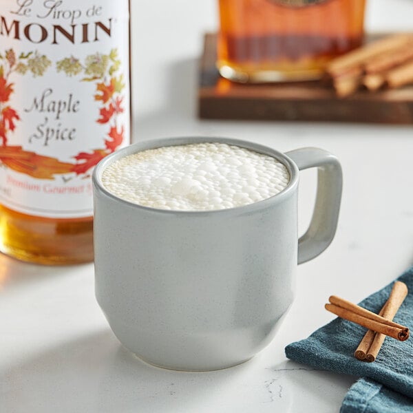 A mug of coffee with foam and cinnamon sticks on a table with a bottle of Monin Premium Maple Spice flavoring syrup.