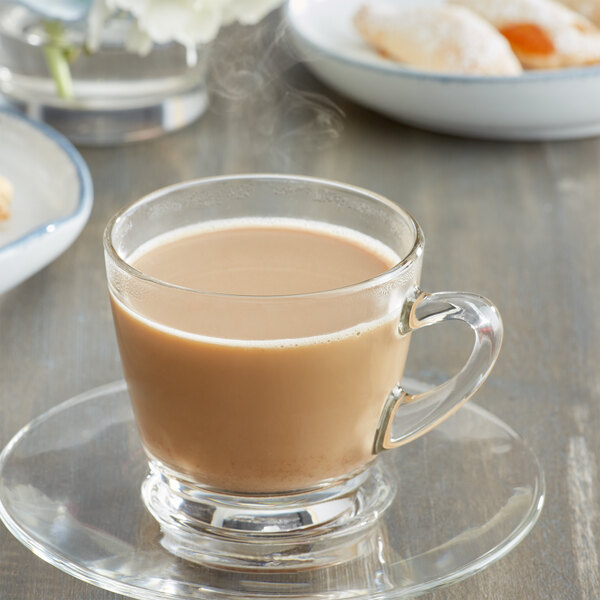 A glass cup of Caffe de Aroma French Vanilla Cappuccino with a spoon on a saucer.