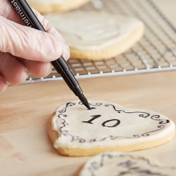 A hand using a Chefmaster black food decorating pen to draw a heart on a cookie.