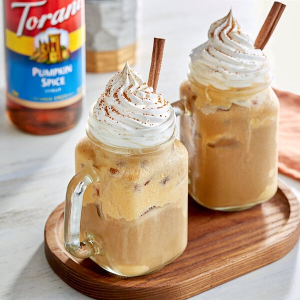 Two mugs of coffee with whipped cream and cinnamon on a table with a bottle of Torani Pumpkin Spice syrup.