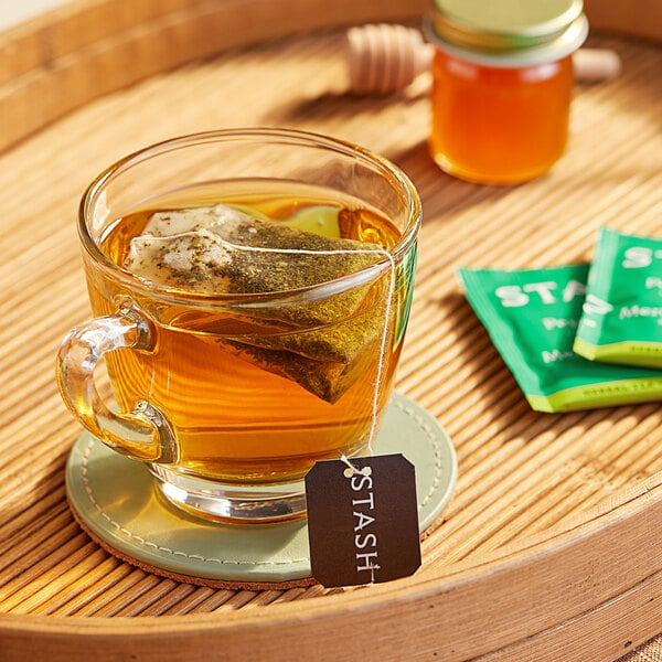 A glass cup of Stash Peppermint Tea with a tea bag in it on a table with a jar of honey.