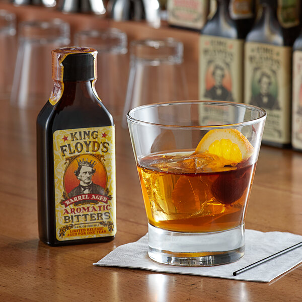 A glass of brown liquid with a slice of orange next to a bottle of King Floyd's Barrel Aged Aromatic Bitters.