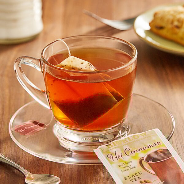 A glass cup of Bigelow hot cinnamon black tea with a tea bag on a saucer.