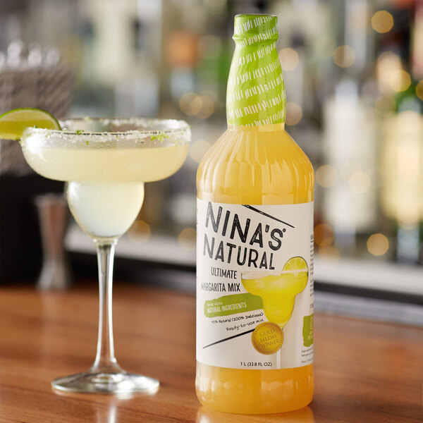 A bottle of Nina's Natural Ultimate Margarita Mix with a glass of margarita with a lime wedge on a table.