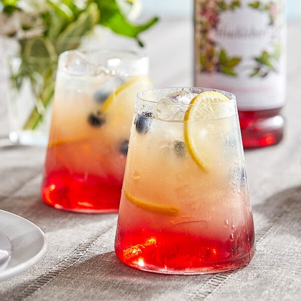 Two glasses of Monin Huckleberry syrup with fruit slices and ice.