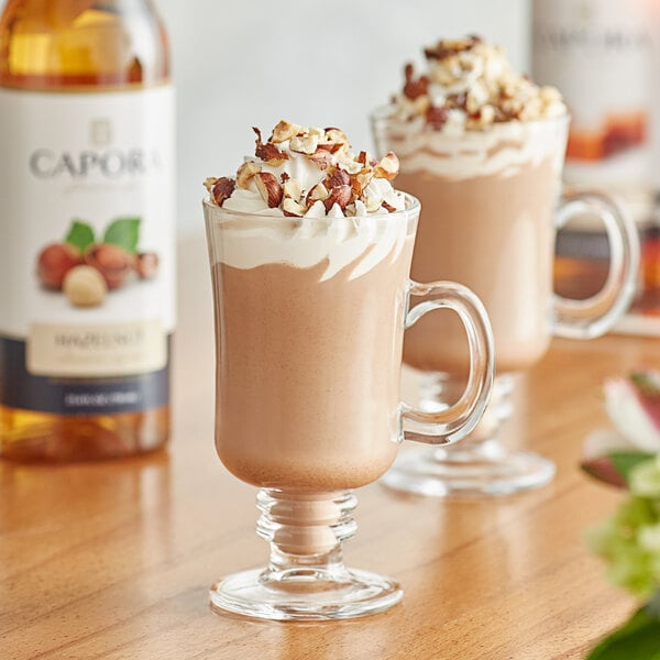 A glass mug of hot chocolate with whipped cream and nuts next to a bottle of Capora Hazelnut Flavoring Syrup.