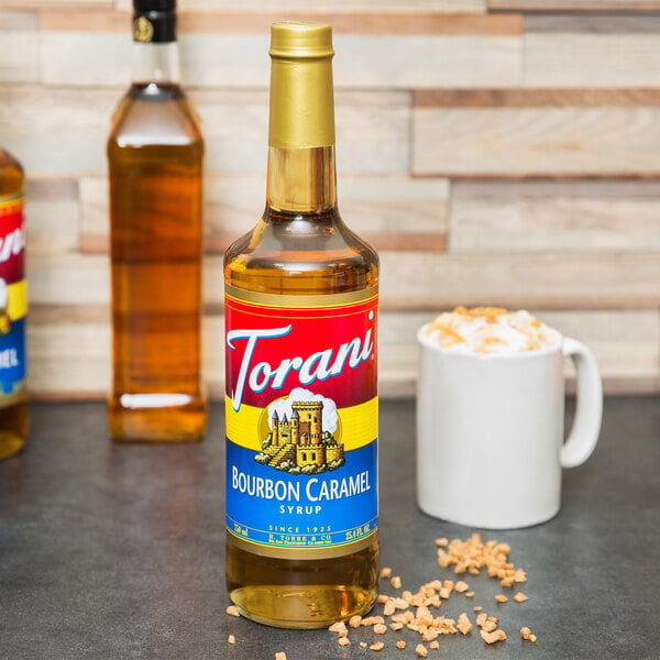 A Torani Bourbon Caramel flavoring syrup bottle on a table next to a white mug with a foamy drink.