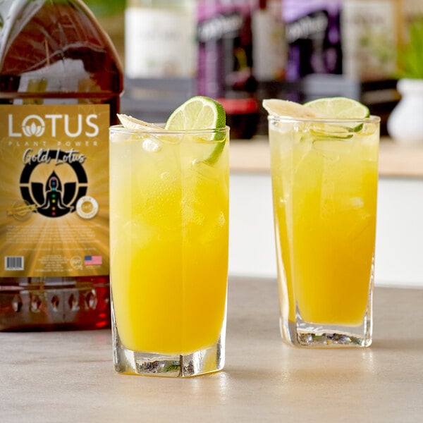 A bottle of Lotus Plant Power Gold Lotus Energy Concentrate next to two glasses of yellow liquid with lime slices.