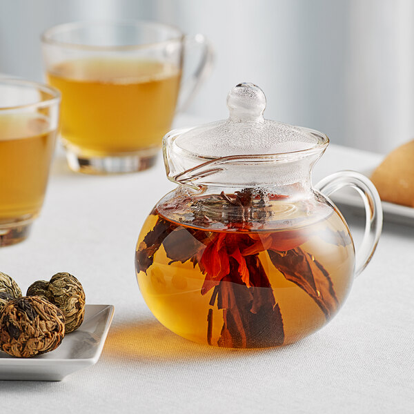 A glass teapot with a flowering tea blossom inside on a white table.