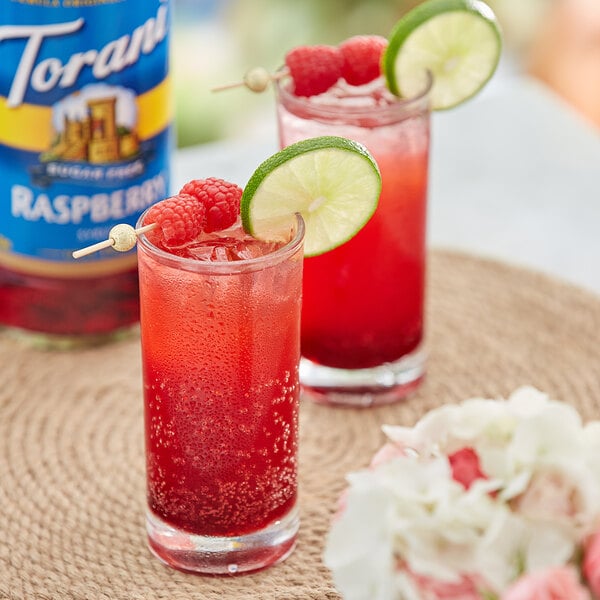 Two glasses of red drinks with lime and raspberry garnishes with a jar of raspberries in the background.