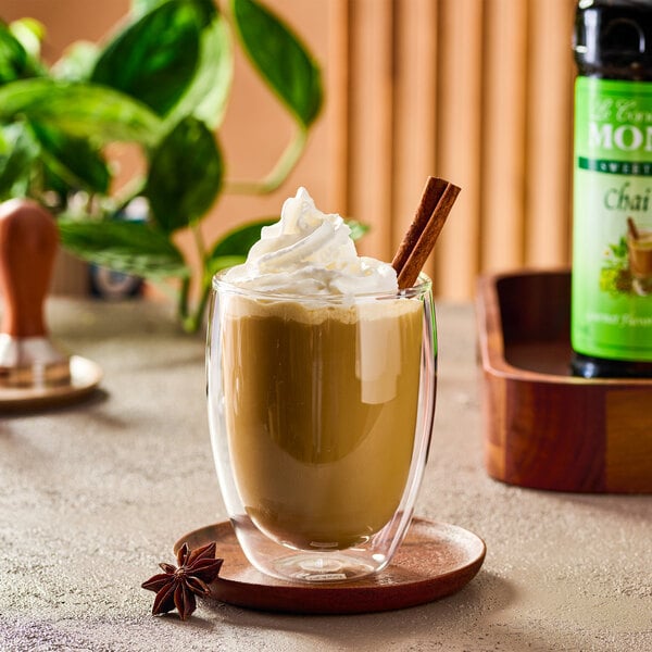 A glass mug of chai tea topped with whipped cream and a cinnamon stick, placed on a wooden saucer with a star anise beside it.