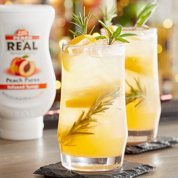 Two glasses of peach puree infused syrup with rosemary sprigs on a table.