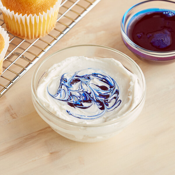 A bowl of white frosting with blue swirls next to a cooling rack with a Chefmaster True Blue Liqua-Gel cupcake.