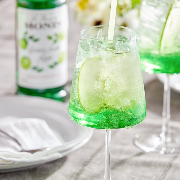 A glass of green liquid with a straw on a table with a bottle of Monin Premium Granny Smith Apple Fruit Syrup.