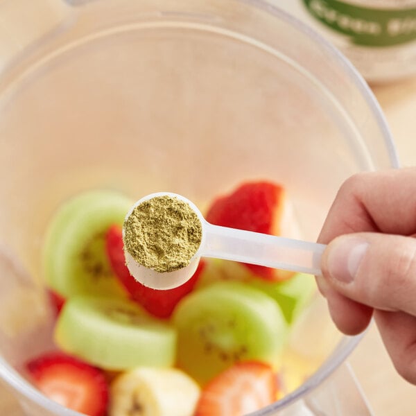 A hand using a measuring spoon to add Add A Scoop Green Blend supplement powder to fruit.