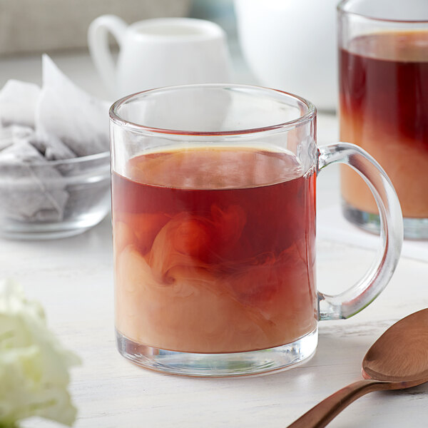 A glass mug of brown Earl Grey tea on a white table with a spoon.