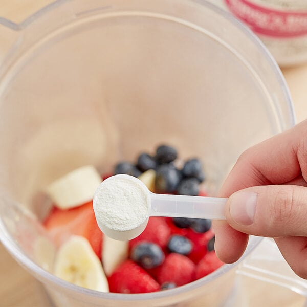 A hand using a scoop to add powder to a blender with fruit.