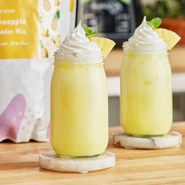 Two glass jars of yellow pineapple smoothie with whipped cream on top.