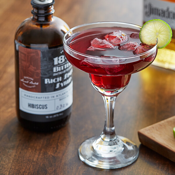 A glass of hibiscus drink with ice and a lime slice next to a bottle of 18.21 Bitters Hibiscus Syrup.