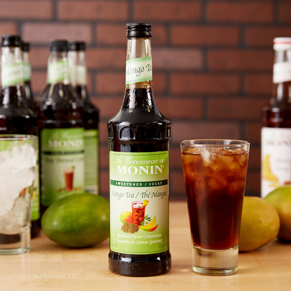 A close-up of a Monin Mango Tea Concentrate bottle with a glass of brown liquid with ice.